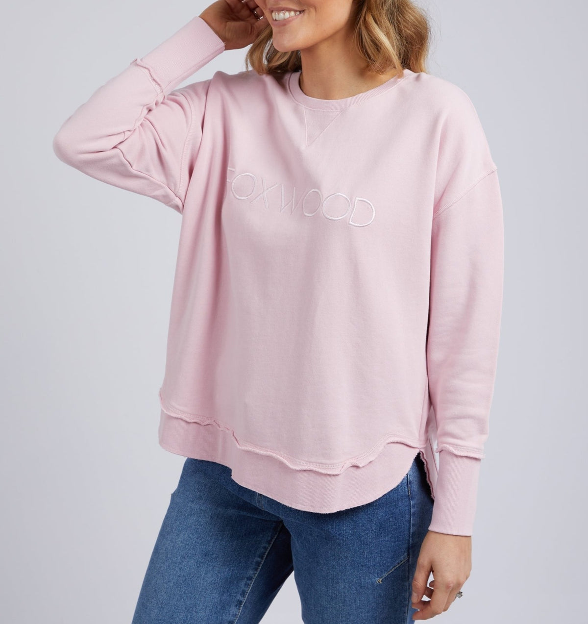 A woman in a relaxed fit, light pink Foxwood Simplified Crew made from 100% cotton with "OXWOOD" embroidered on the front smiles and touches her hair, wearing blue jeans and standing against a plain background.