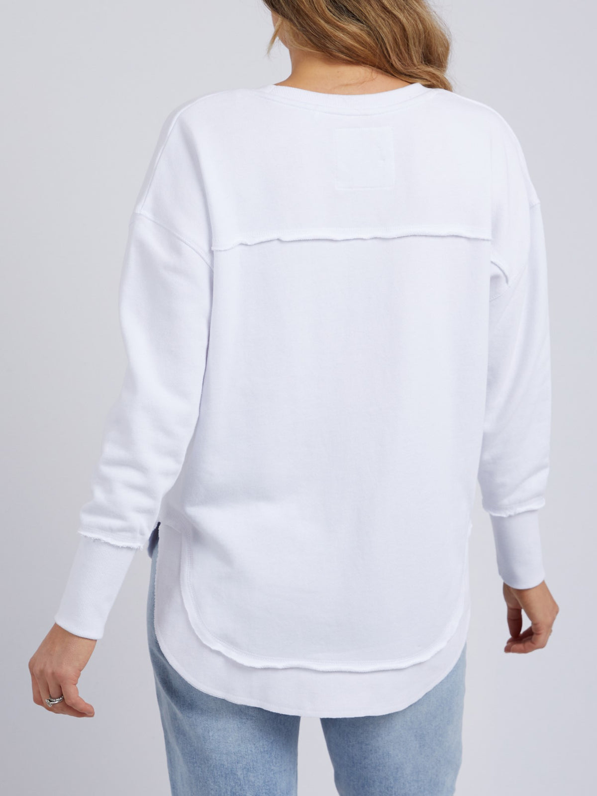 A person with long, wavy hair is shown from behind, wearing a relaxed fit Foxwood Simplified Crew in white with light blue jeans against a plain background.