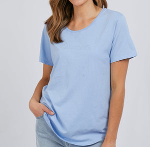 A woman stands casually with one hand in her pocket, wearing a light blue 100% cotton Foxwood Tee with short sleeves and light blue jeansβa simple, effortless style against a plain background.