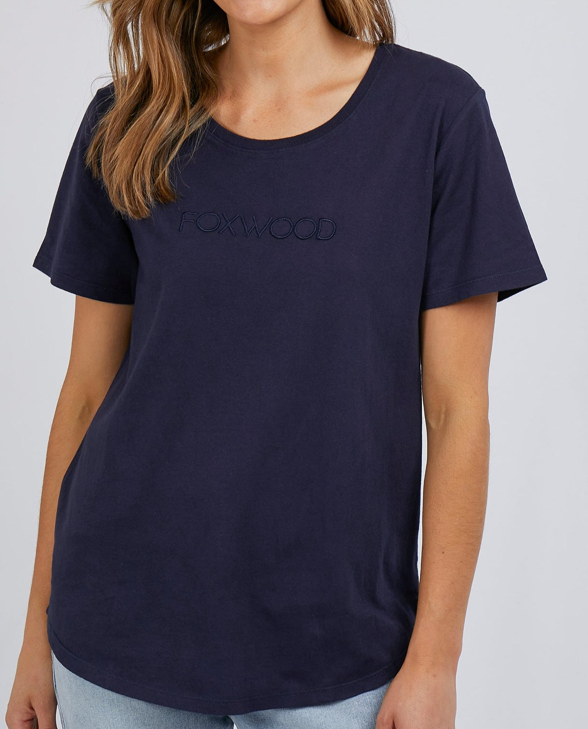 Against a white background, someone models the Foxwood Tee, a 100% cotton dark blue classic with "FOXWOOD" subtly embossed on the chest, perfectly paired with light blue jeans.