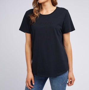 A woman wears the Foxwood Tee, a casual 100% cotton staple paired with blue jeans. The tee features a subtle embossed word, short sleeves, and a round neckline on a neutral gray backdrop. Her face is not visible.