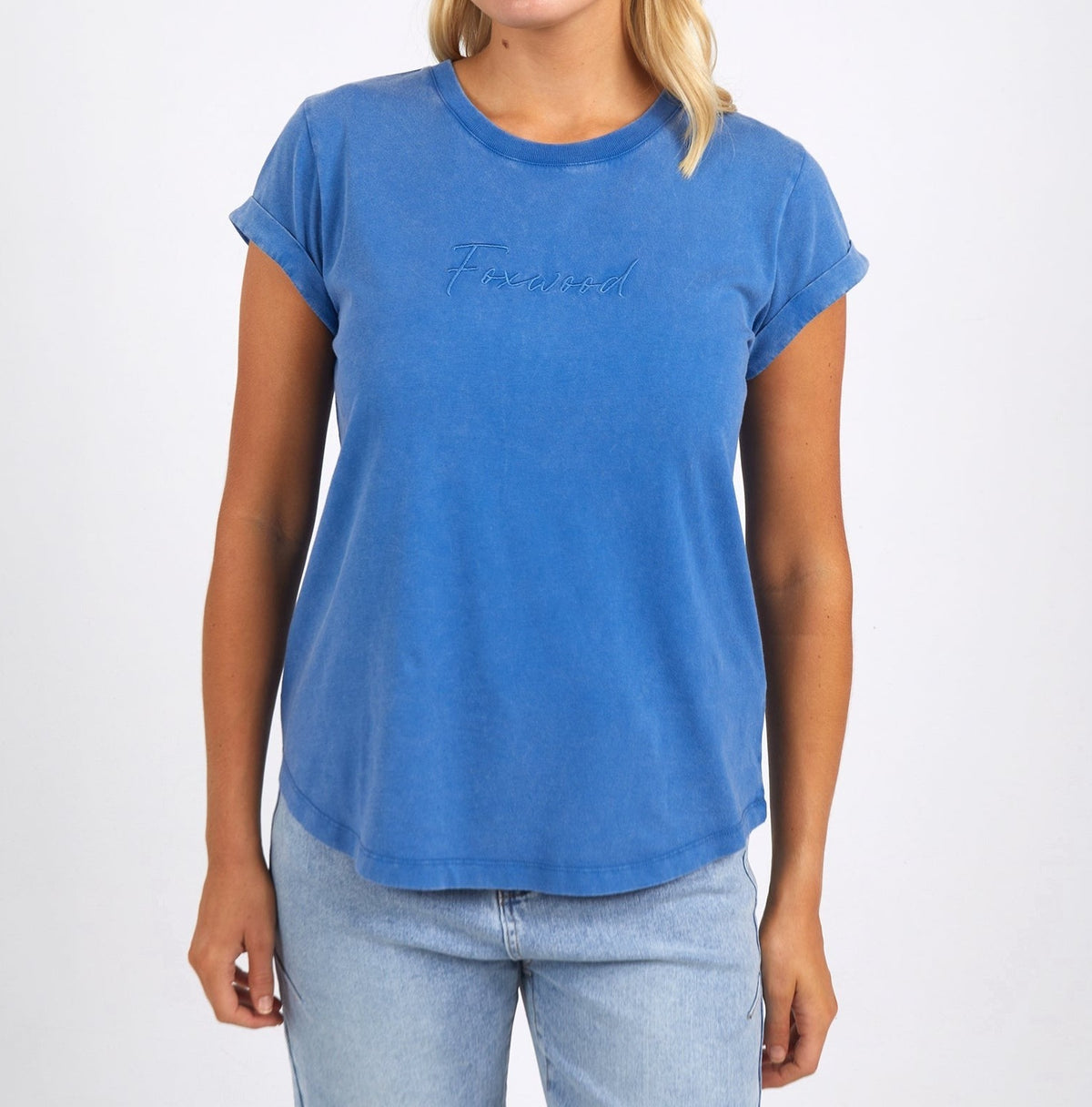 A person models the Foxwood Signature Teeโa blue rolled-sleeve T-shirt with "Friend" embroideryโpaired with light blue jeans. The plain white backdrop highlights its classic style and lightweight, breathable 100% cotton fabric.