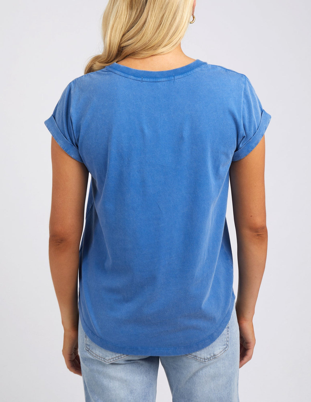 A person with long blond hair is seen from behind wearing the Foxwood Signature Tee, a blue T-shirt made of lightweight breathable fabric with rolled sleeves, paired with light blue jeans. The tee has a rounded hem and is set against a plain white background.
