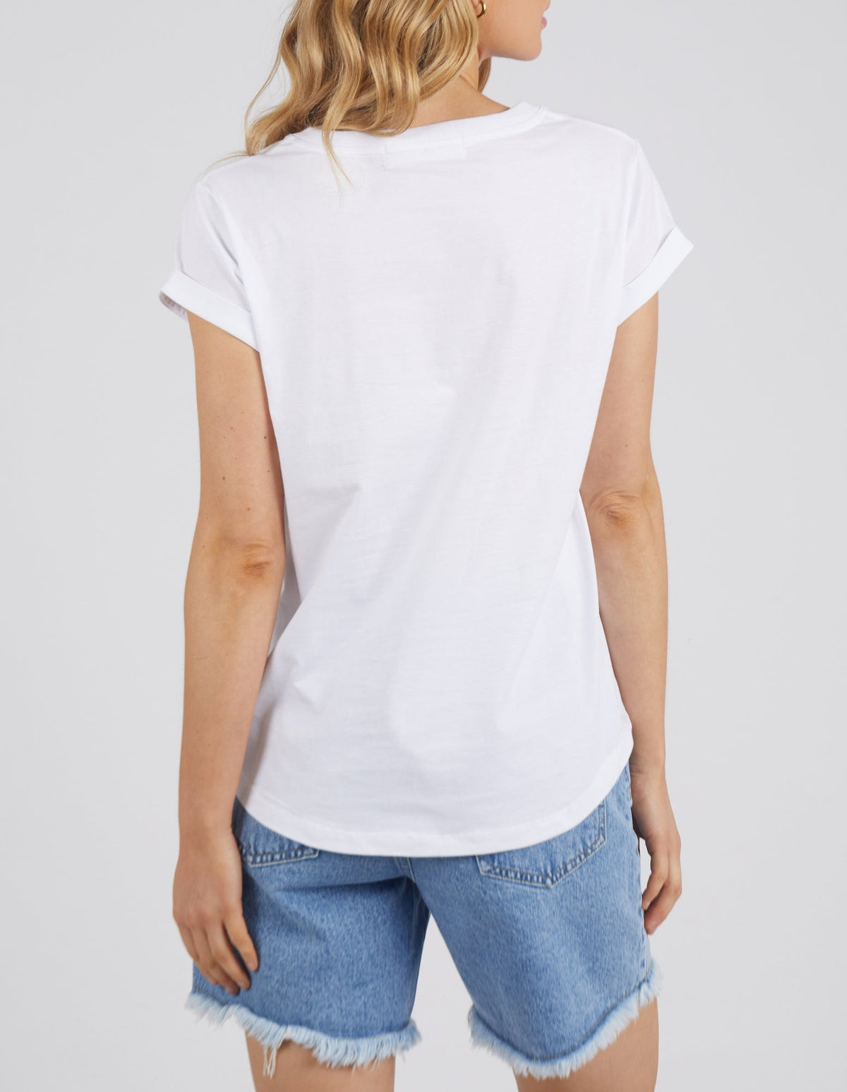 A person with wavy blonde hair is seen from behind, wearing a plain white Foxwood Manly Vee Tee and light blue frayed denim shorts, creating an effortlessly stylish contemporary look against a light background.