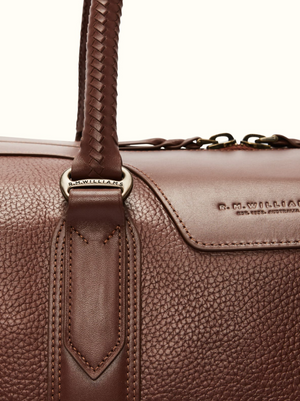 This RM Williams Overnight Bag, made from full-grain leather with a textured surface, features a braided handle, logo-adorned metal ring, and embossed "R.M.WILLIAMS" with small text beneathβideal for all your travel needs.