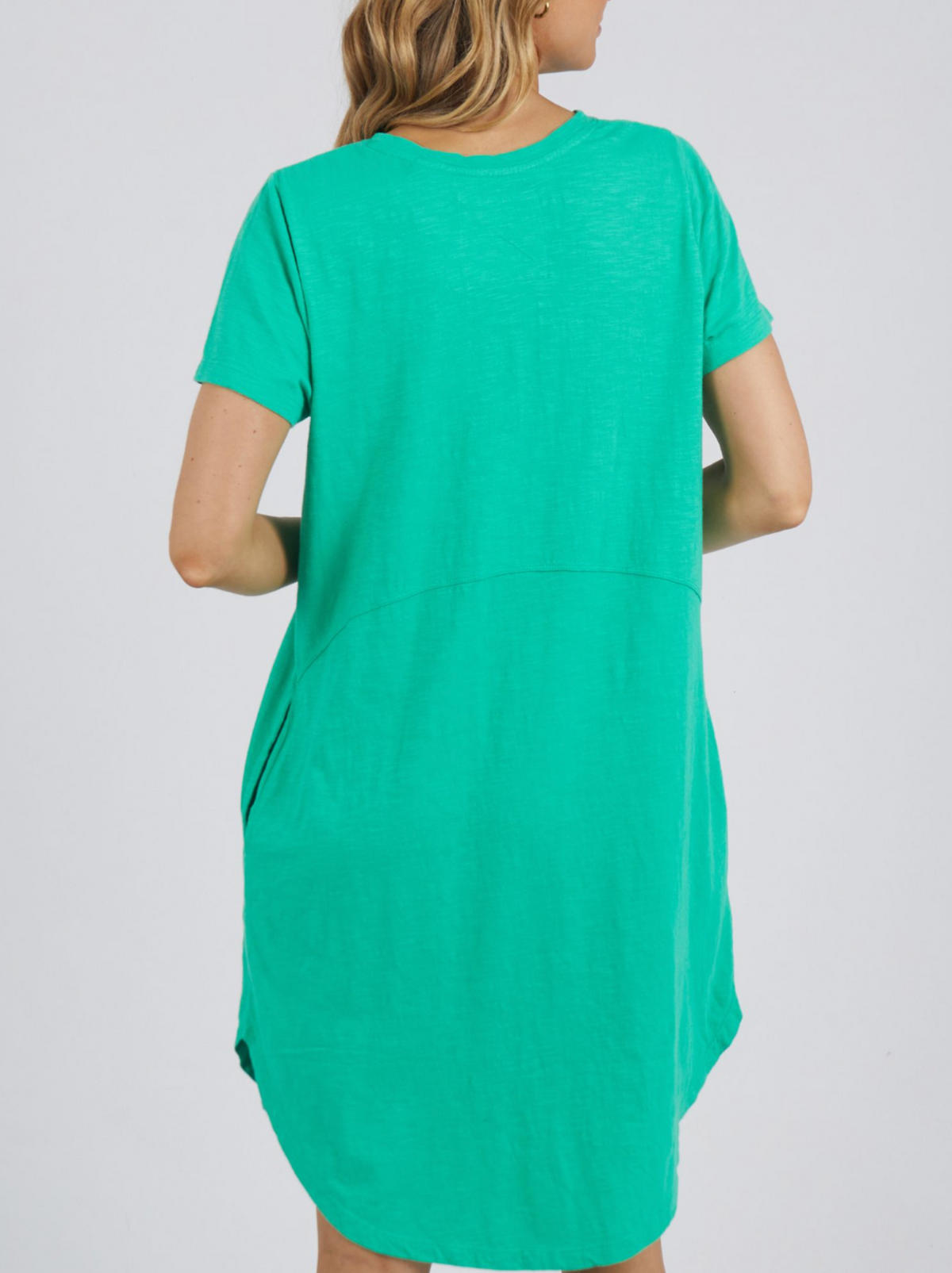 A person is shown from the back wearing a short-sleeved Foxwood Bay Dress in a plain, light background. The dress is 100% cotton jersey, loose-fitting with a slightly curved hem and visible waist seam.