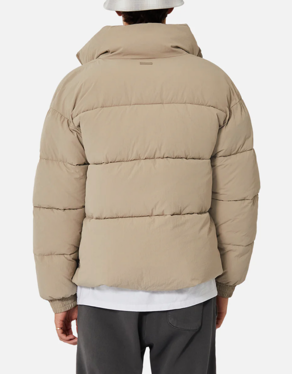 A person dressed in the Industrie The Regent Puffer Jacket, featuring quilted padding, a high collar, and horizontal stitching for added texture, stands with their back facing the camera. They are also wearing dark pants, a white shirt underneath, and a white bucket hat against a plain white background.