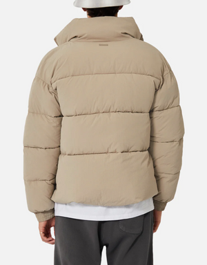 A person dressed in the Industrie The Regent Puffer Jacket, featuring quilted padding, a high collar, and horizontal stitching for added texture, stands with their back facing the camera. They are also wearing dark pants, a white shirt underneath, and a white bucket hat against a plain white background.