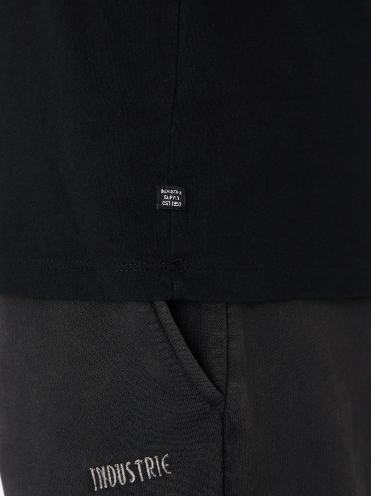 Close-up of a person in black pants with "INDUSTRIE" embroidery and the Industrie The Basic Crew Tee, a 100% cotton Classic Fit with a tag reading "INDUSTRIE SUPPLY EST 2002." Everyday Essential style.