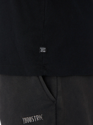 Close-up of a person in black pants with "INDUSTRIE" embroidery and the Industrie The Basic Crew Tee, a 100% cotton Classic Fit with a tag reading "INDUSTRIE SUPPLY EST 2002." Everyday Essential style.