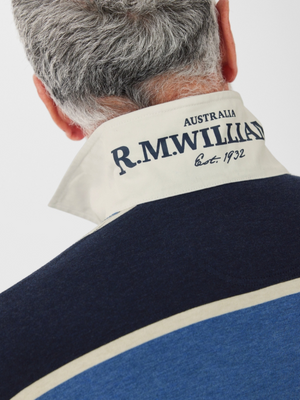 A gray-haired individual wears the RM Williams Tweedale Rugby, featuring a navy and blue striped design with a white upturned collar embroidered with "AUSTRALIA R.M.WILLIAMS est. 1932," crafted in heavyweight jersey knit fabric.