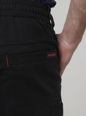 Close-up of a person’s right hip and hand wearing the Hard Yakka Tough Maxx Pant, a durable work pant with an elastic waistband. Visible: black pocket with a red and black "Hard Yakka" label; shirt partly tucked in.