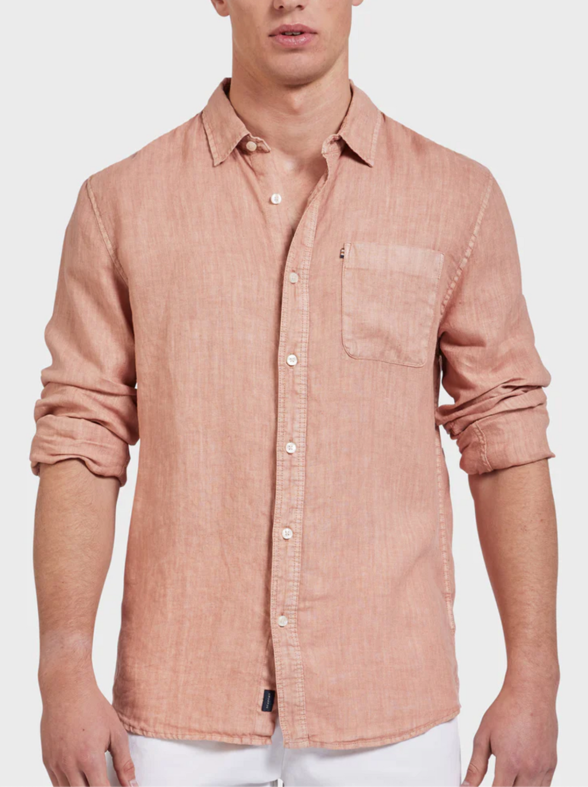 A man wears the Academy Brand Hampton Linen Shirt in light pink with white buttons and a chest pocket, paired with white pants. The shirt’s sleeves are rolled up for a relaxed vibe.