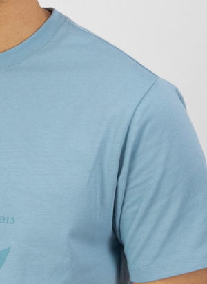 Close-up of a person wearing the Swanndri Original v2 Print T-Shirt in light blue classic fit, showing part of the chest, shoulder, and arm against a plain background. The face is not visible.