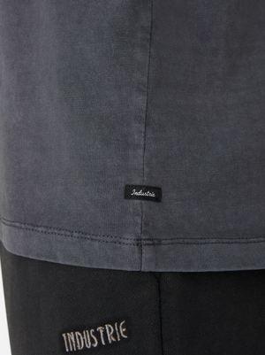 Close-up of a person's side wearing the oversized Industrie The Del Sur Tee in dark gray, with a small black label near the hem. This 100% cotton T-shirt effortlessly pairs with black pants featuring a white-stitched "Industrie" logo near the waistband.