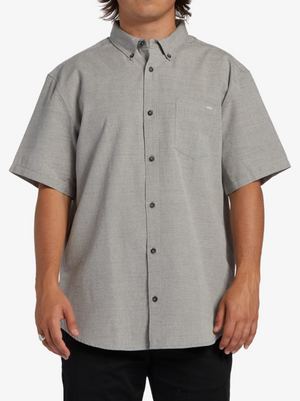 A person wears the Billabong All Day Short Sleeve Shirt in light gray, made from cotton polyester woven fabric, with a collar and chest pocket. Its subtle pattern pairs well with black pants, all on a plain white background.