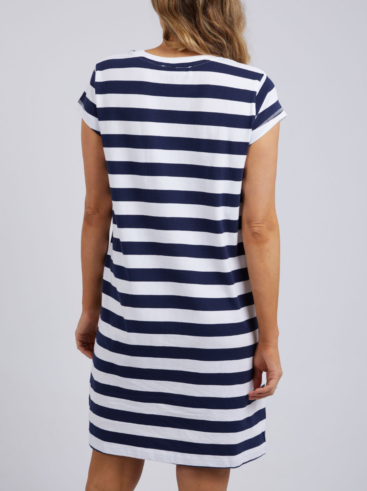 The person wears a Foxwood Signature Stripe Tee Dress in navy blue and white stripes, made of 100% cotton jersey. The knee-length dress has short sleeves, and their long, wavy light brown hair flows down their back.
