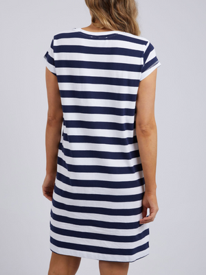 The person wears a Foxwood Signature Stripe Tee Dress in navy blue and white stripes, made of 100% cotton jersey. The knee-length dress has short sleeves, and their long, wavy light brown hair flows down their back.