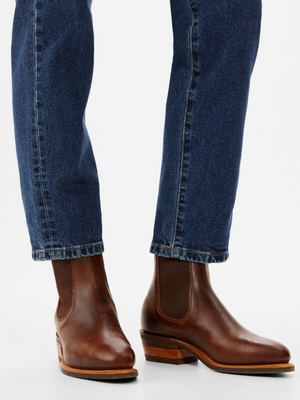 Close-up of a woman in RM Williams Lady Yearling Rubber Sole Boots, dark brown with stacked heels, paired with slightly cropped blue jeans. The plain white background highlights the comfort insole and full view of the boots.