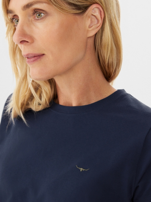 A blonde woman looks to the side while wearing the RM Williams Piccadilly T-Shirt in navy blue cotton jersey, featuring a small longhorn embroidery on the chest. The background is plain and light-colored.