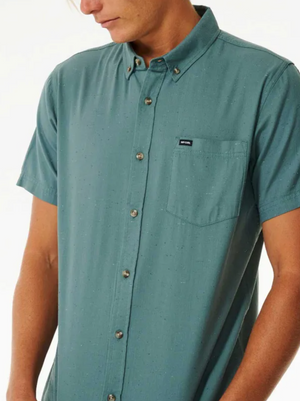 A person wears a Rip Curl Ourtime Short Sleeve Shirt in teal, featuring small speckles, a chest pocket with a logo tag, and buttons. They face slightly downward, highlighting only their upper torso.