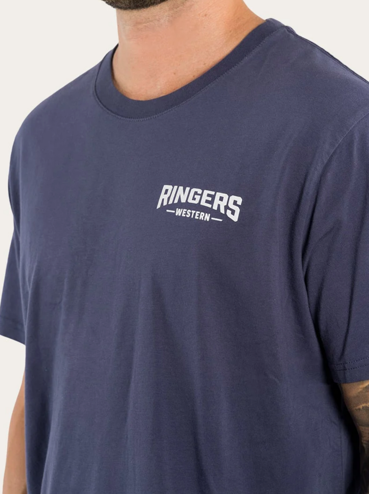 A person wears a blue Ringers Western Squadron Loose Fit T-Shirt, 100% Cotton Jersey, with "RINGERS WESTERN" in white on the left chest. Only the upper torso and part of a tattooed arm are visible.