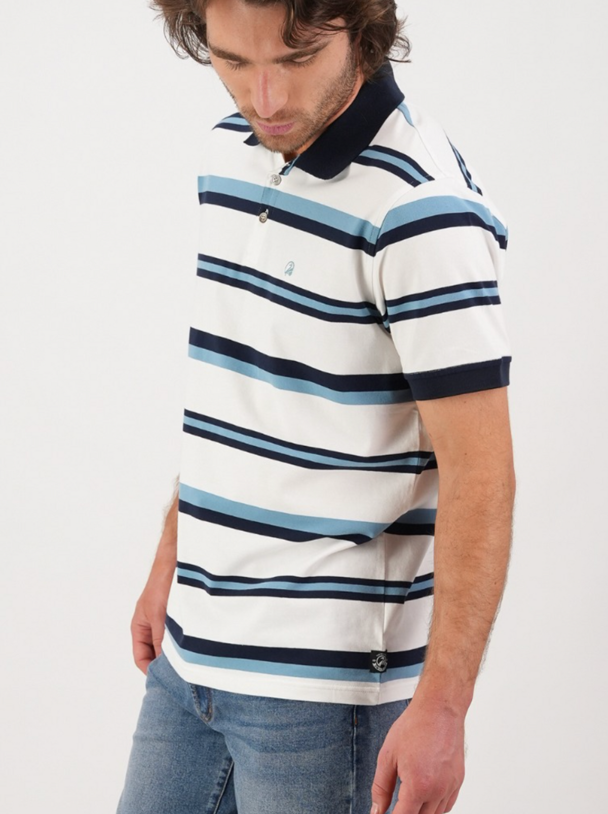 Wearing the Swanndri Winfred Stripe Polo with horizontal blue, navy, and white stripes paired with jeans, a person stands against a plain white background, gazing down with a neutral expression.