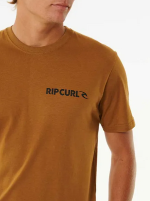 A person wears the Rip Curl Brand Icon Tee in brown, made from 100% cotton. The shirt has a standard fit and features the black "RIP CURL" logo with wave symbol on the left chest. Only their upper body and part of their face are visible.