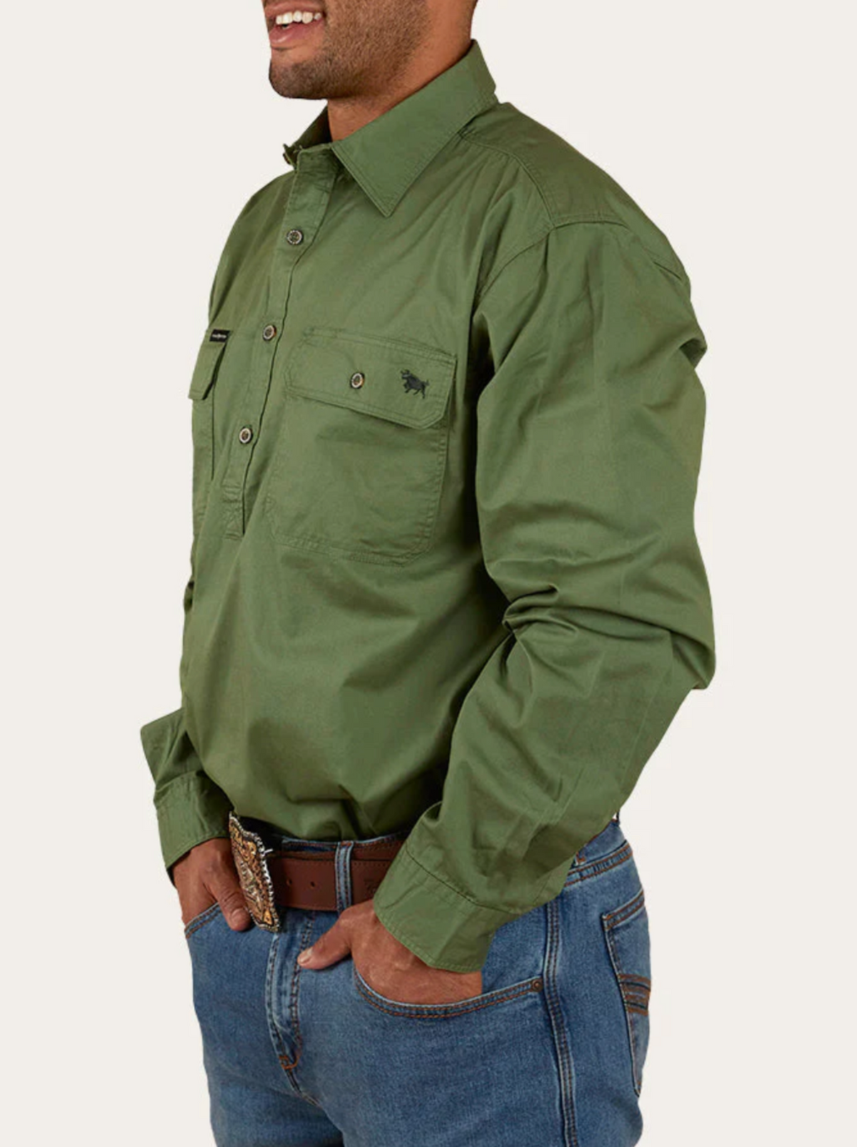 A person is standing sideways with hands in pockets, dressed in a long-sleeved Ringers Western King River Half Button Shirt made of 100% cotton and blue jeans. They are wearing a brown belt with a decorative buckle. The green shirt features a pocket on the right side with a small embroidered logo.