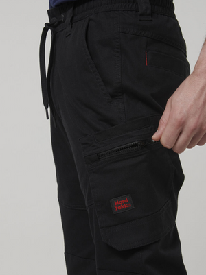 Wearing Hard Yakka Tough Maxx Pant cargo pants with a drawstring waistband, a person unzips a pocket. The pants offer UPF 50+ sun protection and have a red "Hard Yakka" logo patch near the zipper.