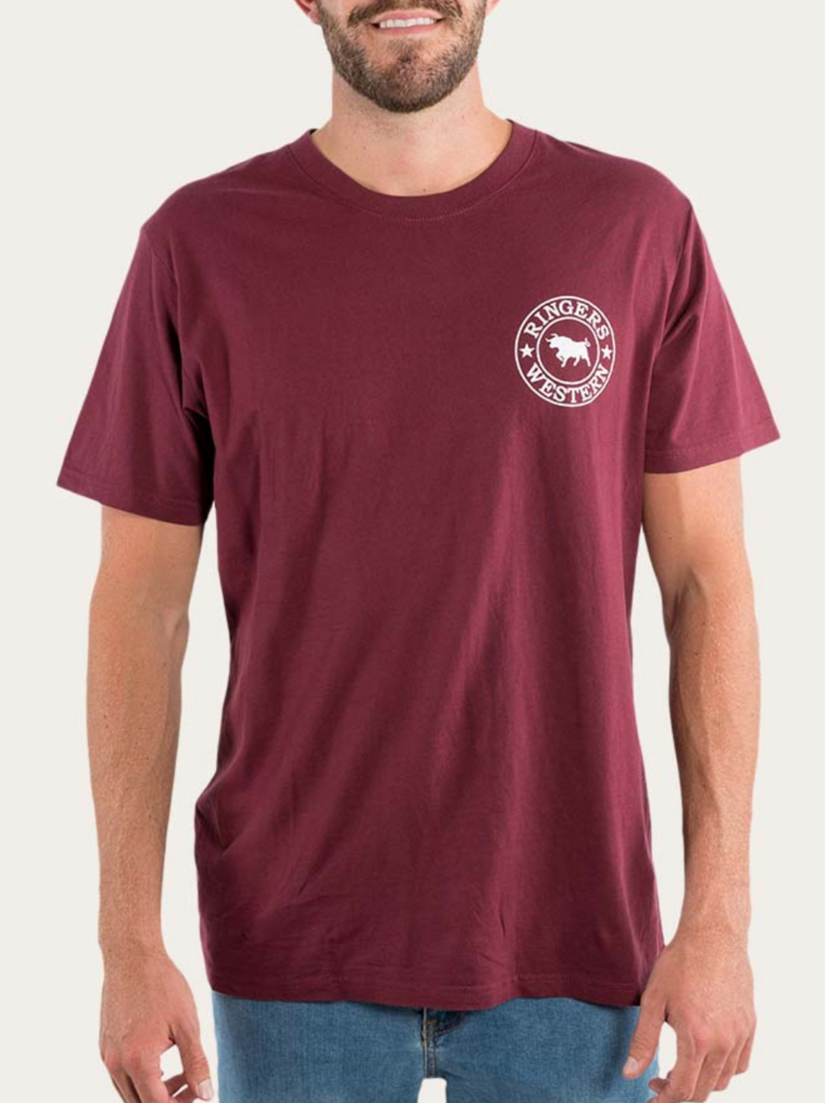 A person is seen wearing a maroon Ringers Western Signature Bull Loose Fit T-Shirt, made from 100% Cotton Jersey for exceptional comfort. The shirt features a small white logo with a horse and the text "Ringers Western" on the upper left chest area. They are also dressed in light blue jeans against a plain, light-colored background.