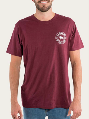 A person is seen wearing a maroon Ringers Western Signature Bull Loose Fit T-Shirt, made from 100% Cotton Jersey for exceptional comfort. The shirt features a small white logo with a horse and the text "Ringers Western" on the upper left chest area. They are also dressed in light blue jeans against a plain, light-colored background.