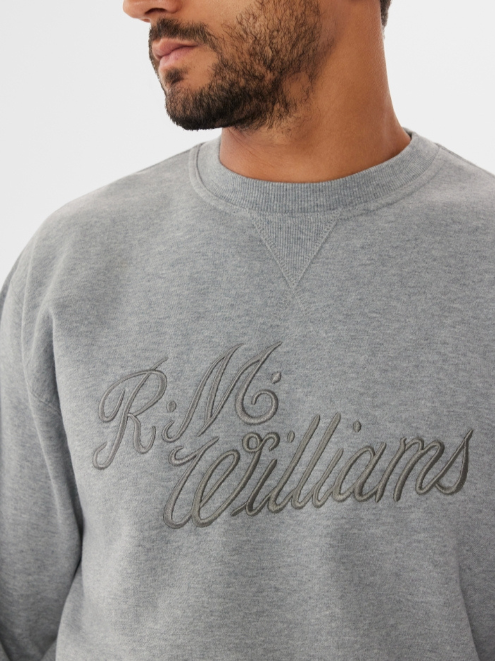 A person with short dark hair and facial hair stands against a plain backdrop wearing the RM Williams R.M.W Script Crew Neck, a gray jumper stylishly embroidered on the front, enhancing their casual yet classic look.