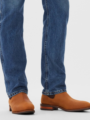 A person wearing slightly faded blue jeans and light brown RM Williams Comfort Craftsman Boots stands against a plain background. The nubuck leather boots showcase their signature quality with a low heel and rounded toe.