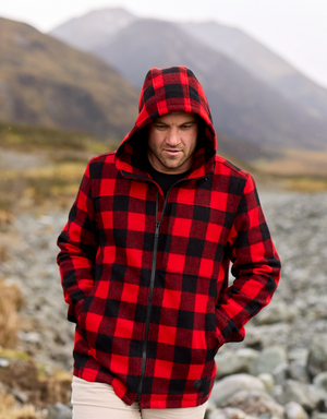 A man wearing a Swanndri Hudson Hoody in red and black plaid stands outdoors in a mountainous landscape. Made from 100% New Zealand wool and featuring a YKK zip, he has his hands in his pockets and looks down slightly, with a rocky path and rugged terrain in the background.