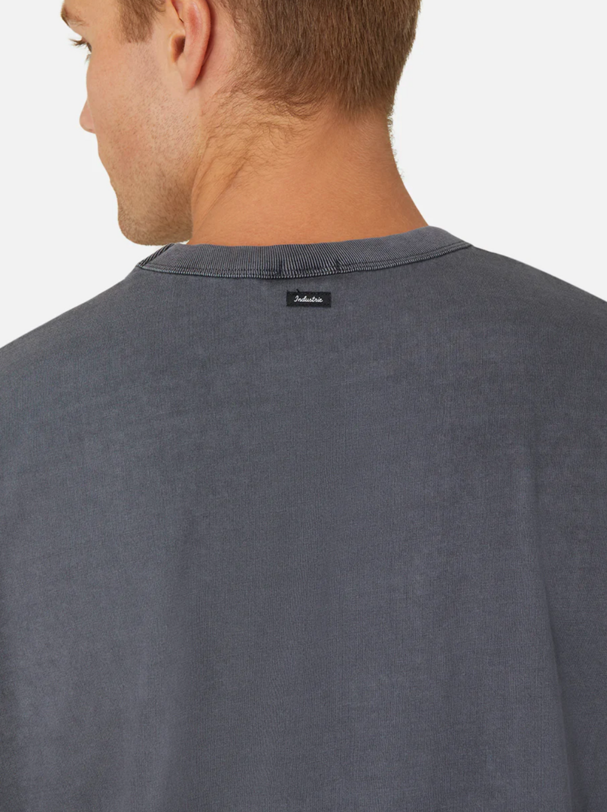 A person wearing an oversized fit, dark gray shirt with a small black label on the back. Made of 100% cotton, the Industrie The Del Sur Tee is captured from behind, highlighting the upper back and neck against a plain white background.