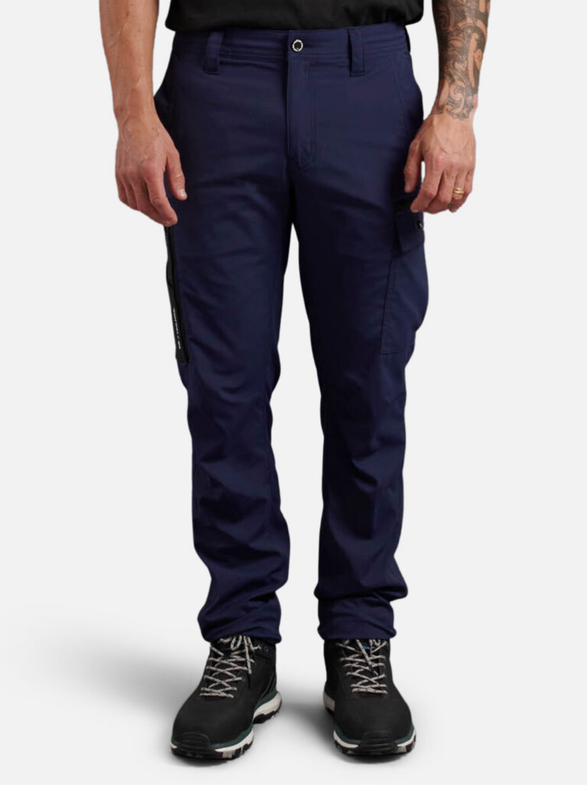 A person in navy King Gee Trademark Cargo Pant stands against a white backdrop, wearing black hiking boots. The pants, made of durable ripstop fabric, have a left thigh pocket and a silver-buckled belt. A tattoo decorates the right arm.
