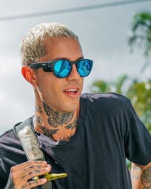 A tattooed individual with short, light hair wears SafeStyle Cruisers Matte Black Frame Mirror Blue Polarised Lens sunglasses and a black T-shirt. They hold a metallic shoulder strap and a smartphone, with palm trees and a cloudy sky in the background.