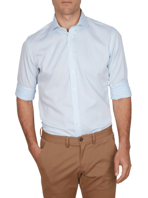 A man wearing the Abelard Non Iron Cotton Twill Slim Fit Shirt in light blue with rolled-up sleeves and tan pants stands with his hands in his pockets against a white background.