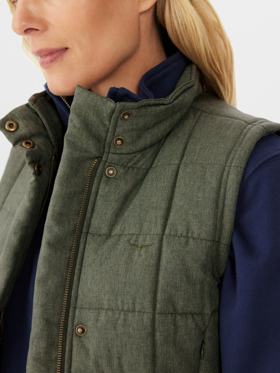 A blonde woman wears the RM Williams Wilpena Creek Vest, a quilted olive green recycled polyester-cotton blend with metal buttons and zipper, over a navy zip-up top. Her face is partially visible in profile, emphasizing functional style.