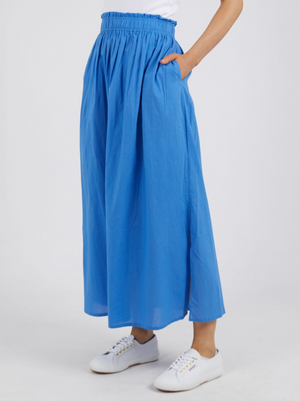 A person is wearing a bright blue, ankle-length Foxwood Charli Skirt with an elastic waistband and pockets. Made of 100% cotton, it pairs perfectly with white sneakers. The background is plain white, creating a simple and clean look.