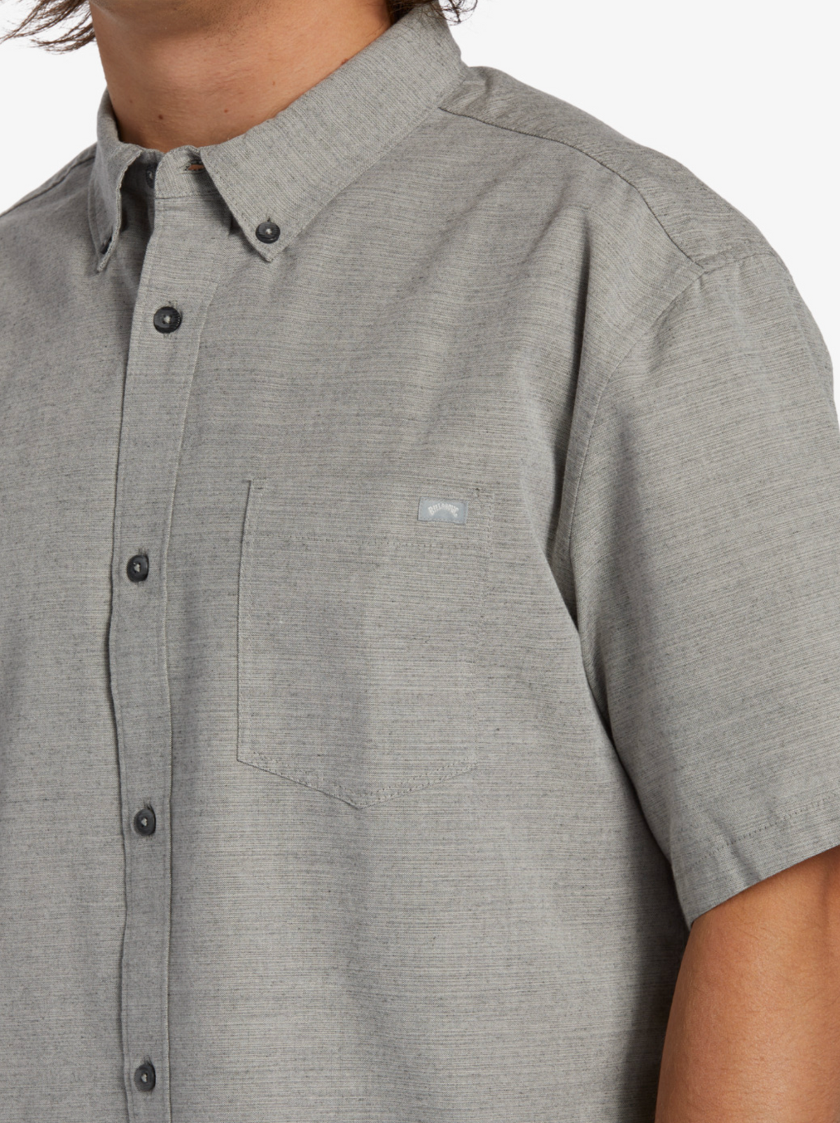 A person is wearing the classic Billabong All Day Short Sleeve Shirt in gray, crafted from woven cotton-polyester fabric. It features dark buttons and a pocket with a subtle logo, with the head partially out of frame, drawing attention to the shirt's timeless design.