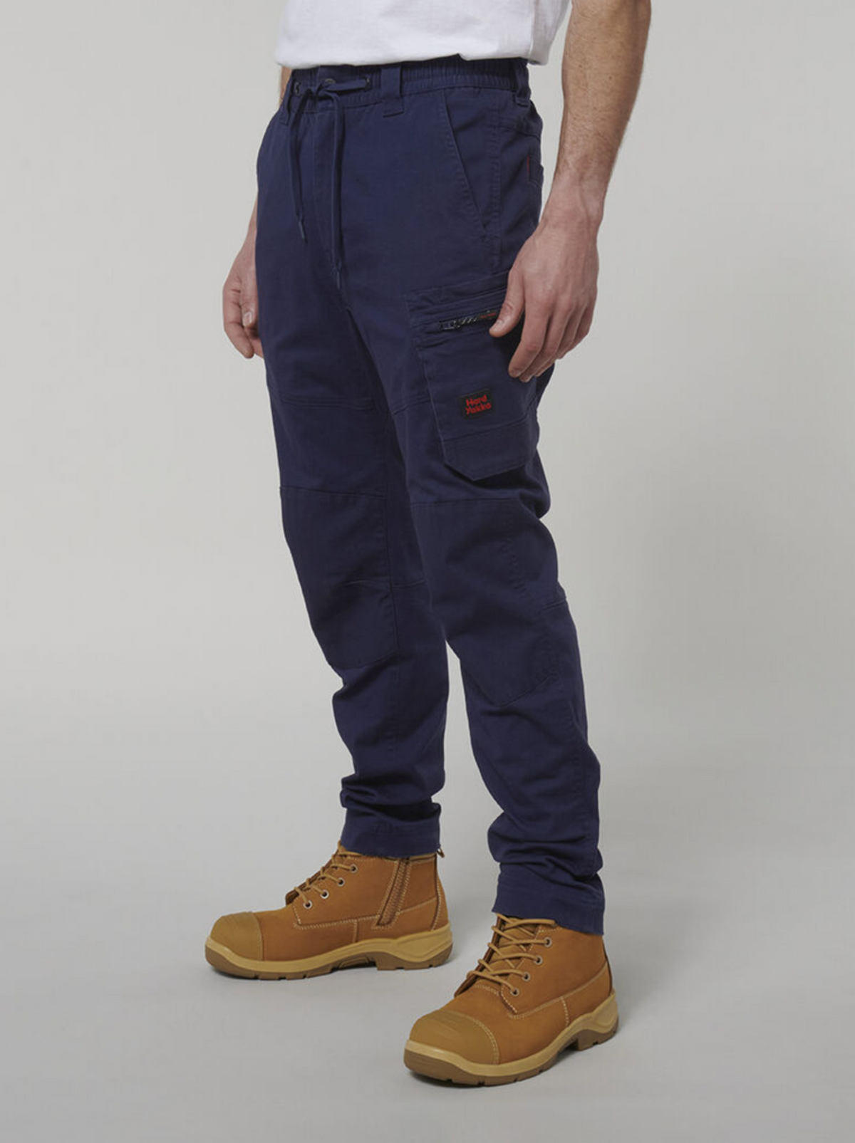 A person dressed in Hard Yakka Tough Maxx Pants in dark blue, featuring a drawstring and multiple pockets, paired with tan work boots and a white T-shirt. The plain gray background highlights their practical style perfectly.