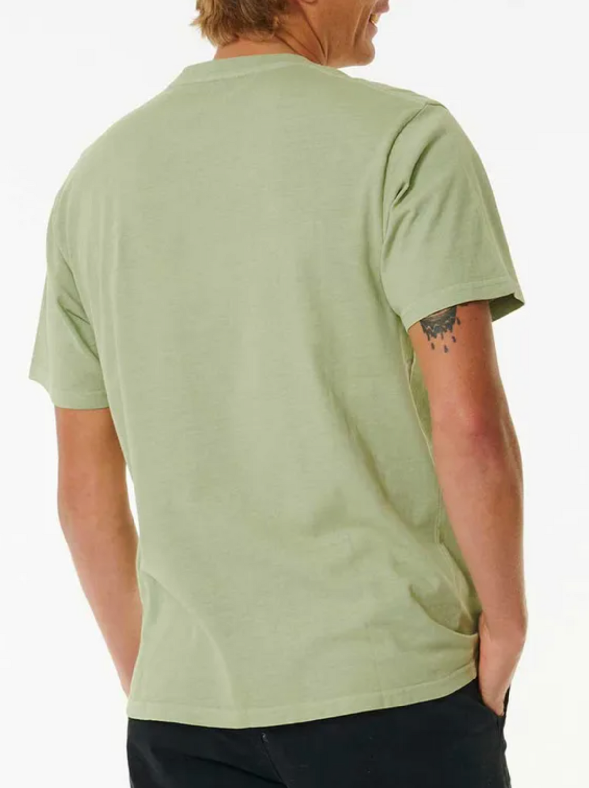 A person is seen from behind wearing a relaxed fit light green Rip Curl Plain Wash Tee and black pants, one hand in a pocket, with a small tattoo visible on their upper right arm.