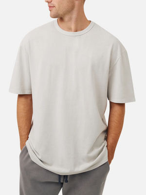 A man wears an oversized light gray Industrie The Del Sur Tee (100% cotton) and dark gray sweatpants, standing with hands in pockets and looking to the side against a plain white background.