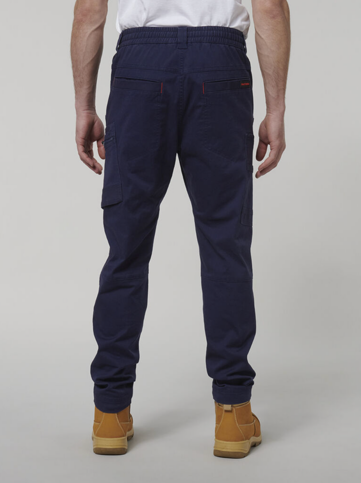 A person in Hard Yakka Tough Maxx Pants, navy blue cargo work pants with back and side pockets, is standing with their back to the camera. They wear tan work boots against a plain light gray background, paired with a white shirt.