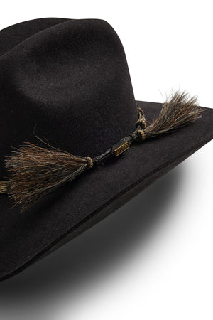 A close-up of the Akubra Rough Rider hat in black felt, featuring a Cattleman crown, decorative braided band with brown and black tassels, a metallic tag, all set against a white background.