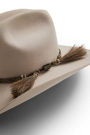 The Akubra Rough Rider is a beige, wide-brimmed felt hat with a braided band, decorative tassels, and a "RUGGED" metal tag—crafted for durability and perfect for any adventure.