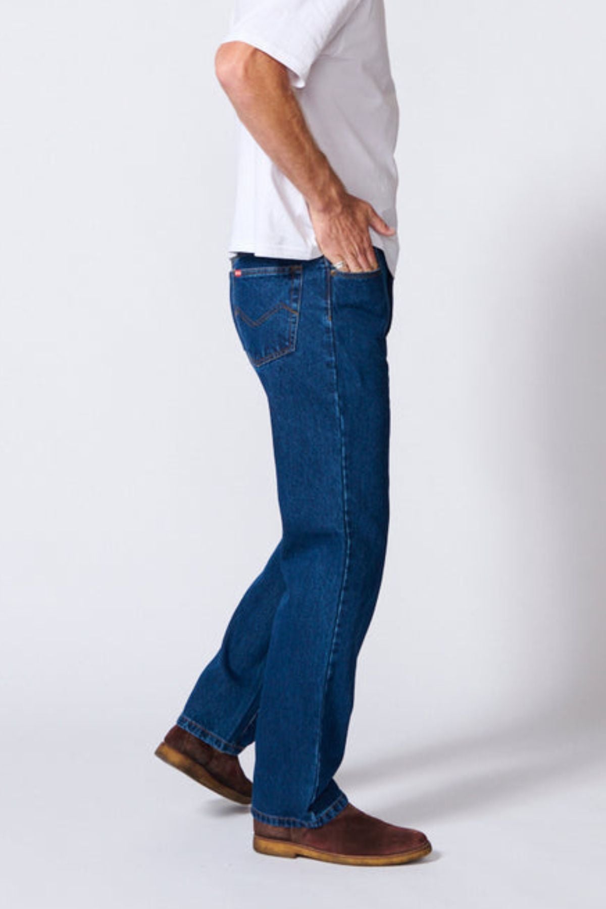 A person in a white short-sleeve shirt, brown shoes, and AMCO Rigid Regular Fit Jean made from 100% cotton denim stands sideways with one hand in their pocket against a neutral background.