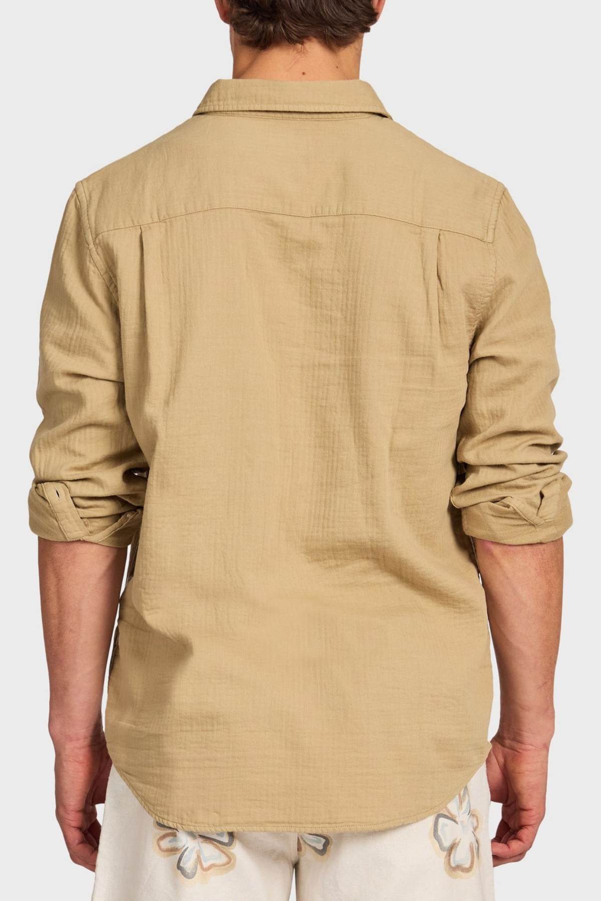 A man is seen from behind wearing the Academy Brand Harry Double Cloth Shirt, a light brown men’s casual shirt with rolled-up sleeves, paired with light floral shorts against a plain white background.